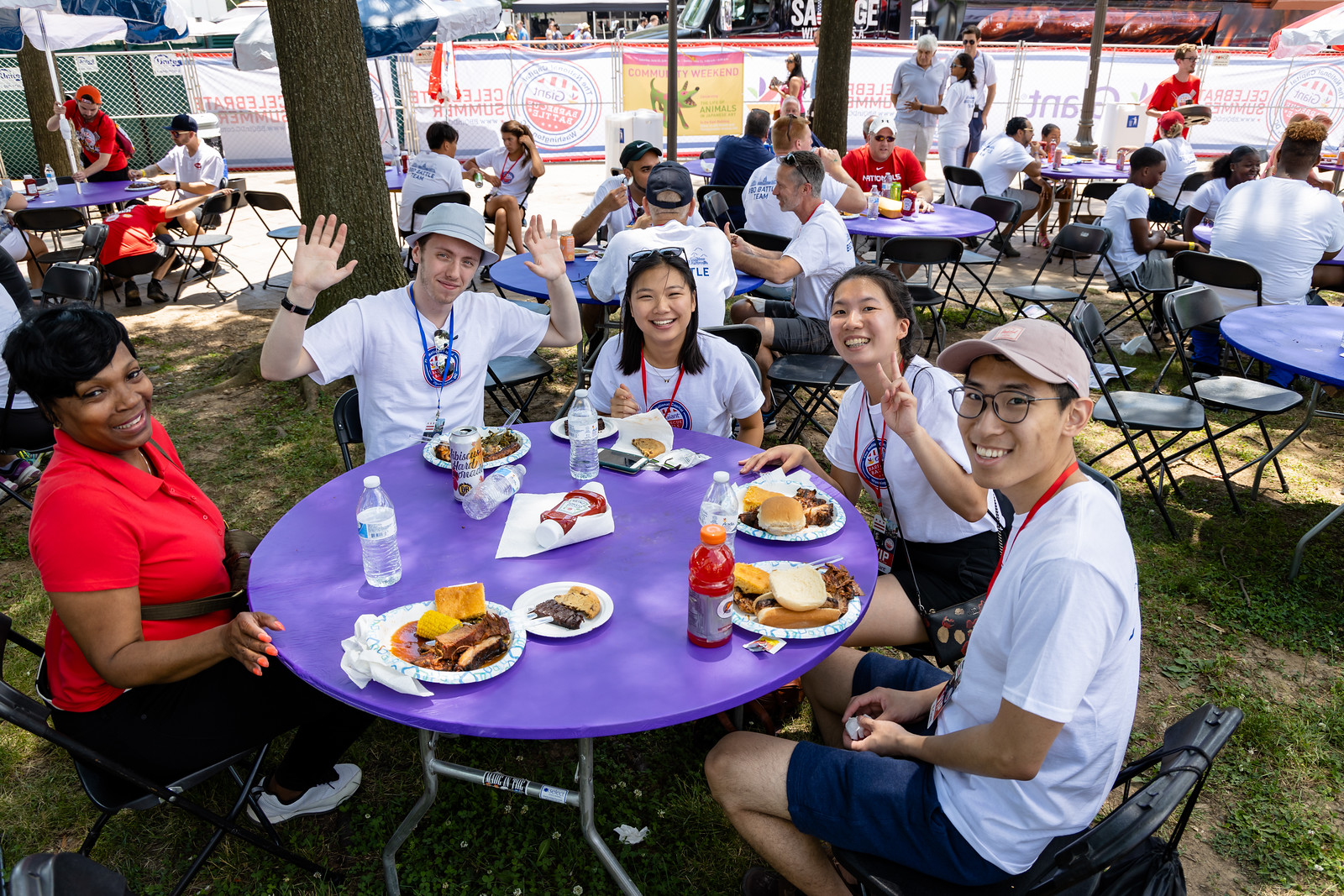 Casual Dining at the Event: A group of six diverse people, smiling and waving, seated around a table enjoying barbecue. 