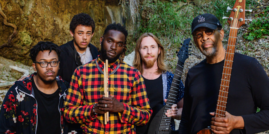 Five musicians posing together, holding instruments, in an outdoor setting.