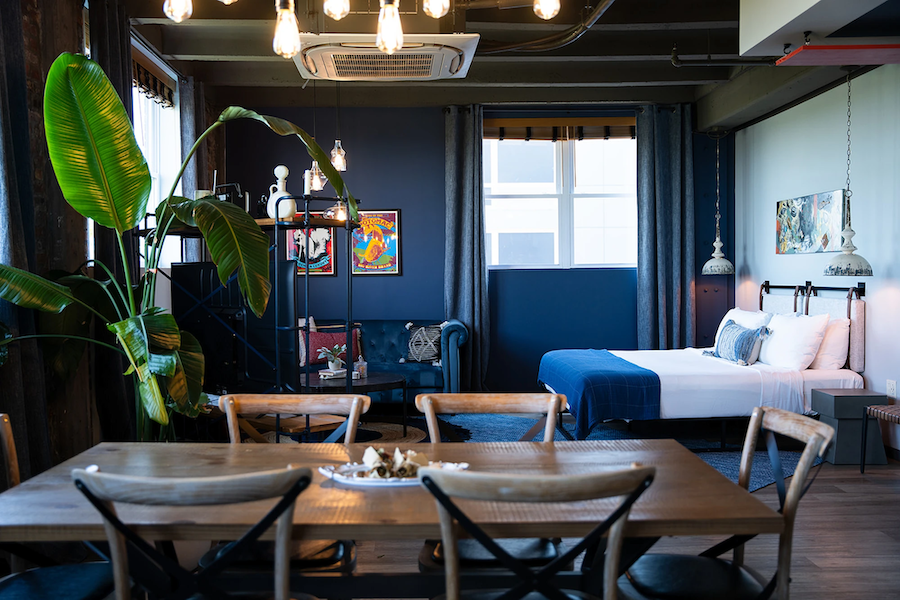 Cozy hotel suite with a dining table, large potted plant, blue sofa, and bed with blue bedding. Dark blue walls and framed artwork add color.