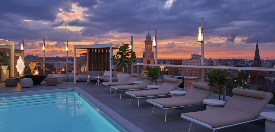 A rooftop pool area with modern lounge chairs and ambient lighting overlooks a cityscape at dusk, with a colorful sky and prominent church tower in the background.