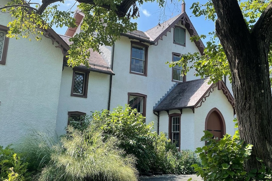 A historic white house with brown trim and gothic-style architecture, surrounded by lush greenery and partially shaded by a large tree.