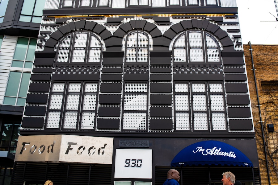 The exterior of The Atlantis, a black and white building with arched windows and geometric patterns on the facade, located at 930. Two people are conversing on the sidewalk in front of the entrance, with a sign above the door that reads 'The Atlantis' in white lettering on a blue awning.