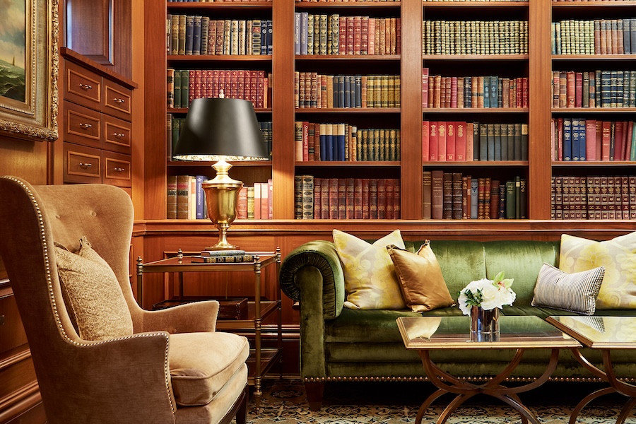 A cozy library with a green velvet sofa, surrounded by bookshelves filled with colorful books. A reading lamp and armchair complete the inviting atmosphere.