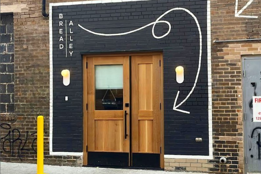 The exterior entrance of Bread Alley, featuring a black brick wall with a white, hand-drawn swirl and arrow pointing towards the wooden double doors. The words "BREAD ALLEY" are vertically aligned on the left side of the entrance, with small wall lights on either side of the door.