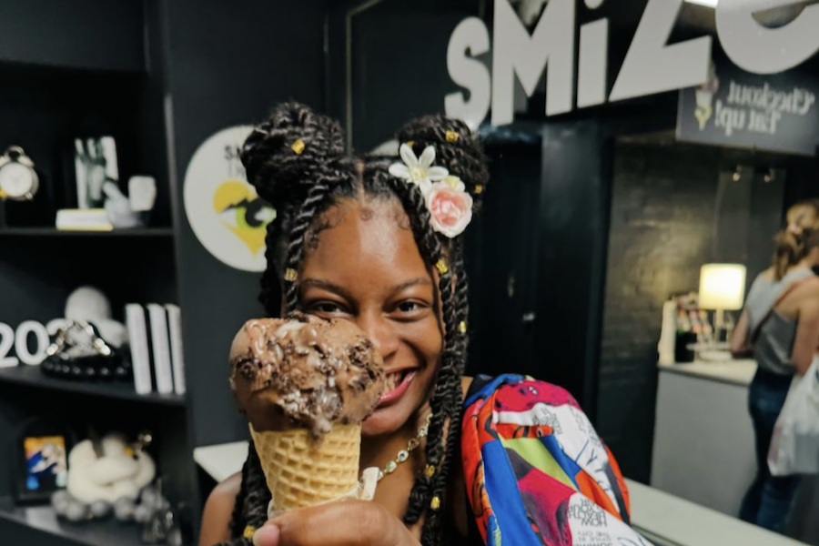 A young girl with braided hair decorated with flowers smiles while holding a waffle cone with a large scoop of chocolate ice cream. She stands inside a shop with the word "SMiZE" visible on the wall behind her. The background includes shelves with various items and other customers.