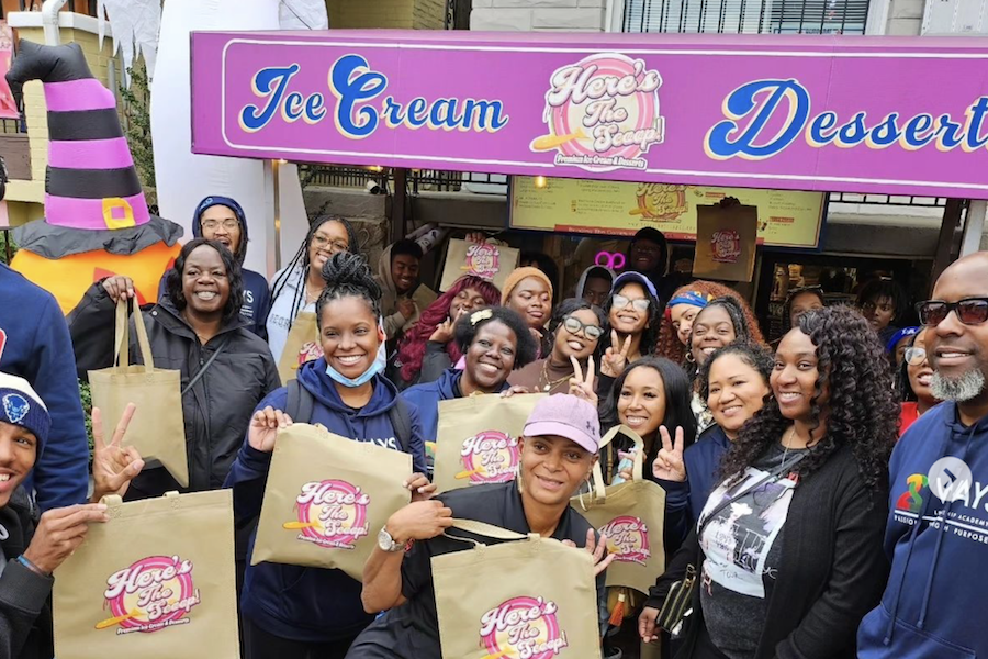 A smiling group holding "Here's The Scoop!" tote bags poses in front of a pink and purple ice cream shop sign. The group is cheerful, with some making peace signs.