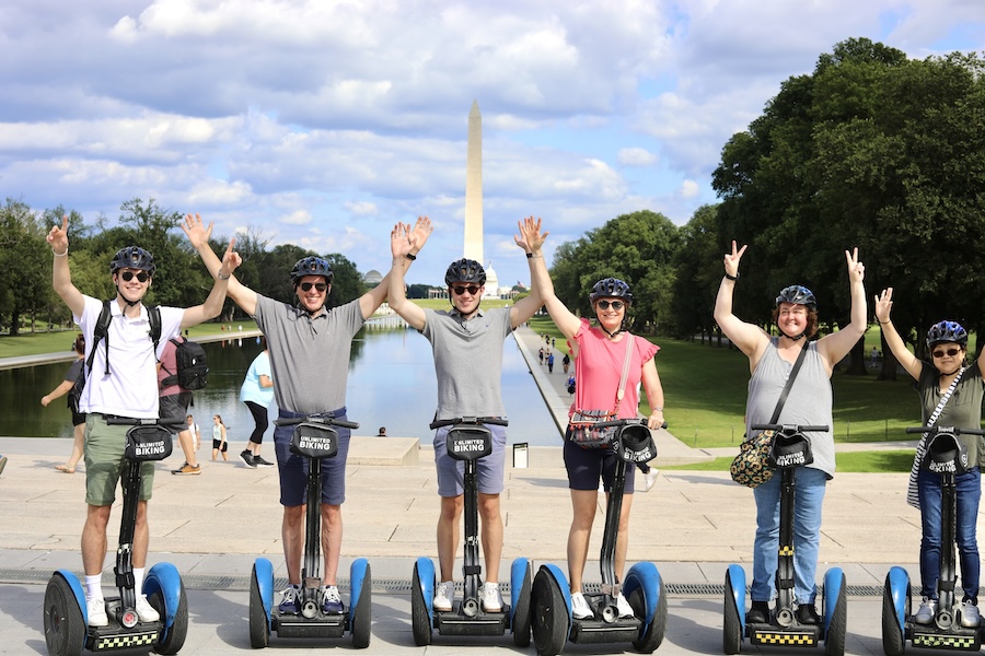 A group of people riding Segways, wearing helmets, and waving in front of the Washington Monument on a bright day.