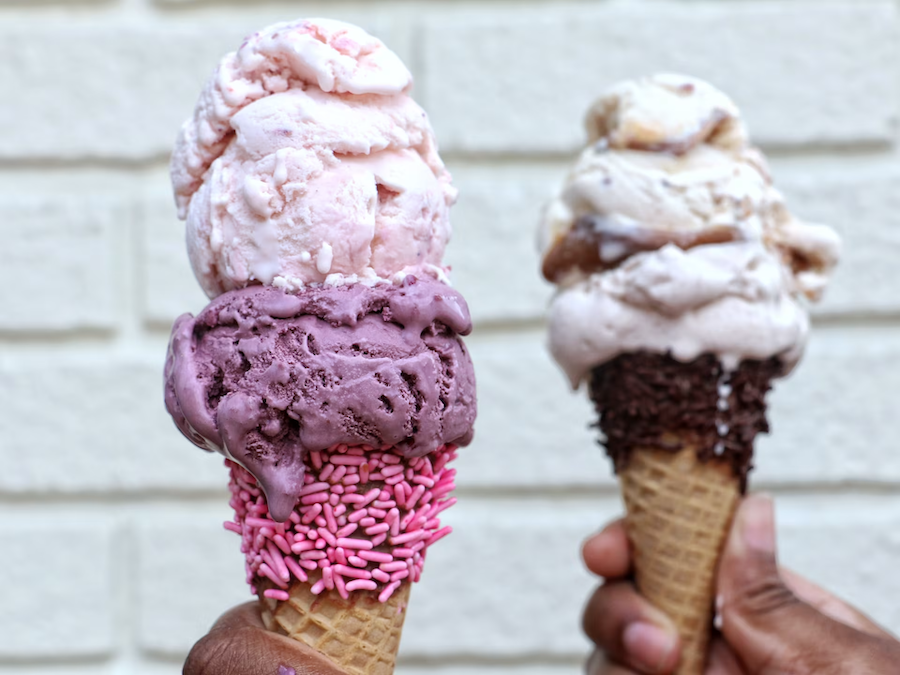  Two hands holding ice cream cones topped with double scoops. The left cone features pink and purple ice cream with pink sprinkles, while the right cone has scoops of brown and white ice cream with chocolate sprinkles, against a white brick background.