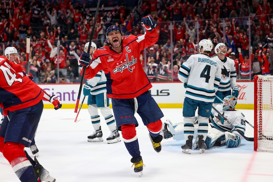 A Washington Capitals player celebrates after scoring a goal, as teammates and fans in red cheer enthusiastically, while the opposing team looks dejected by the net.