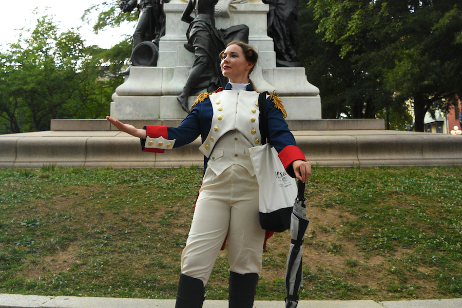 A woman dressed in a historical military uniform, resembling the attire of Marquis de Lafayette, poses confidently in front of a monument. She stands on a grassy area with trees in the background, holding a tote bag and an umbrella.