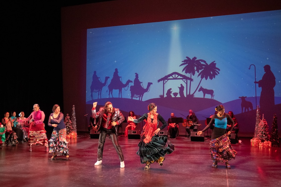 A group of dancers performing a vibrant flamenco routine on stage, with a nativity scene backdrop projected behind them. The dancers wear colorful, traditional outfits, and musicians are seated in the background.