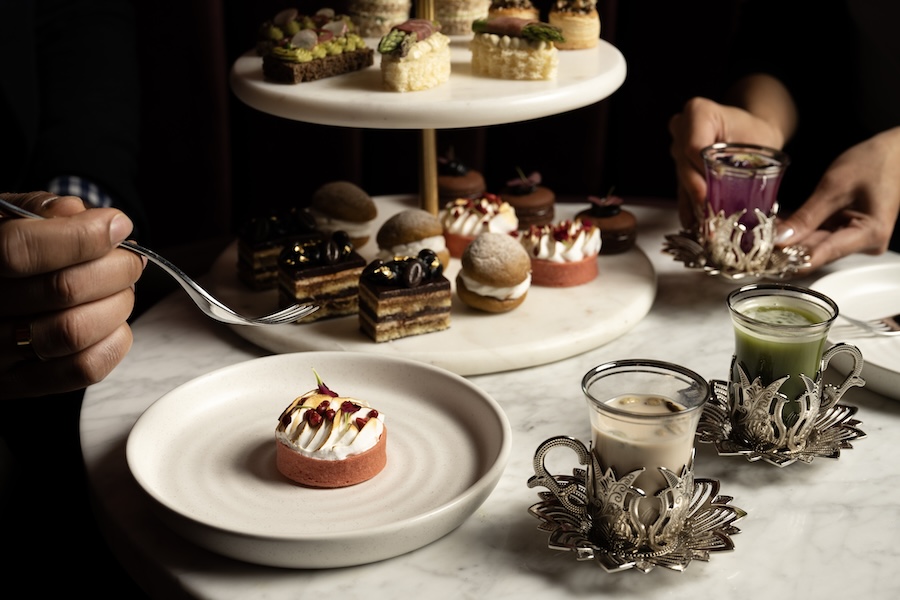 An elegant display of high-tea desserts and vibrant drinks in ornate silver holders, with hands interacting with the treats.