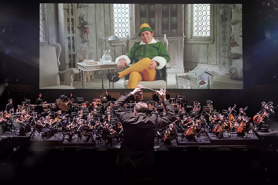 An orchestra performs on stage with a conductor in the foreground, while a large screen in the background displays a scene from the movie 'Elf,' featuring Buddy the Elf sitting in a chair. The performance combines live music with the film.