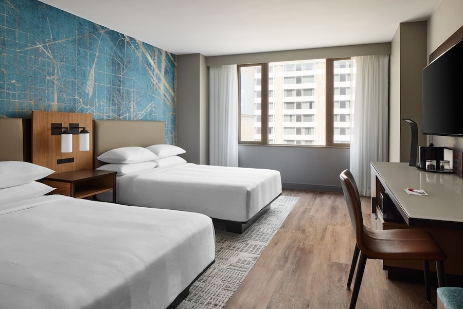 A well-lit hotel room featuring two neatly made queen beds, a large window with sheer curtains, and a modern desk and chair, all set against a textured blue accent wall.