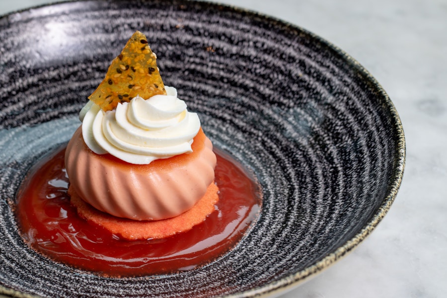 A beautifully presented dessert sits on a textured black plate, featuring a light pink mousse-like dome topped with whipped cream and a honeycomb garnish, resting in a pool of bright red sauce.