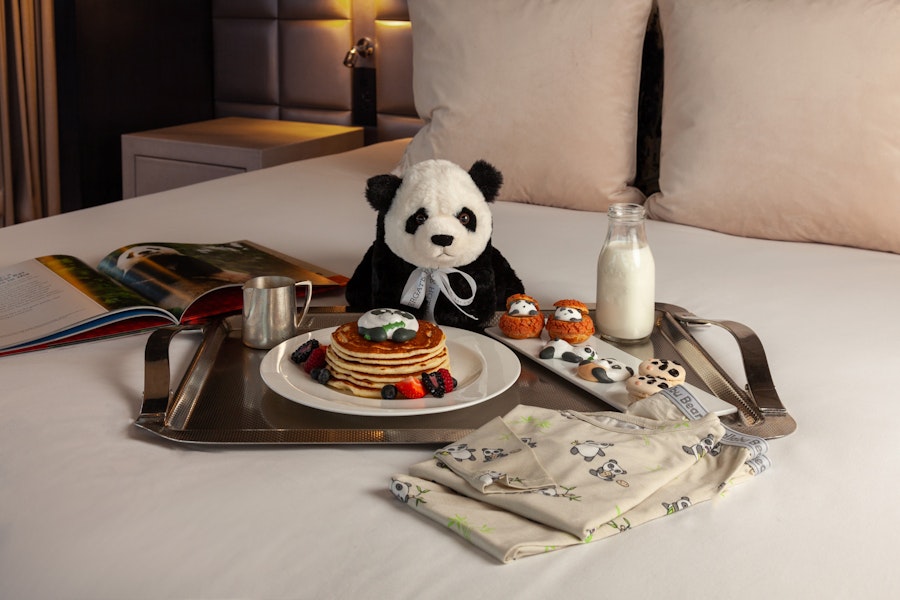 Stuffed animal panda sitting on a crisp, white hotel bed with panda-themed treats.