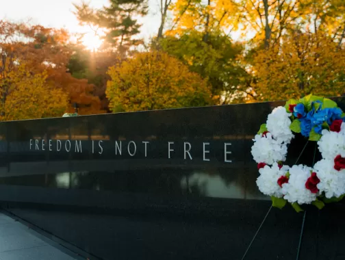 A solemn scene at a memorial, featuring a stone wall engraved with the words "FREEDOM IS NOT FREE." A wreath of red, white, and blue flowers rests against the wall, with autumn trees and the warm glow of a setting sun in the background.