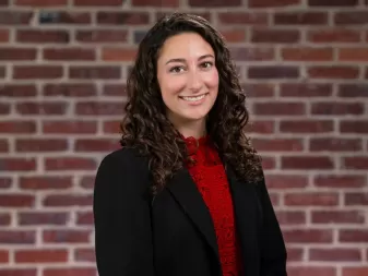 A woman with curly brown hair, wearing a black blazer over a red lace top, stands in front of a brick wall.